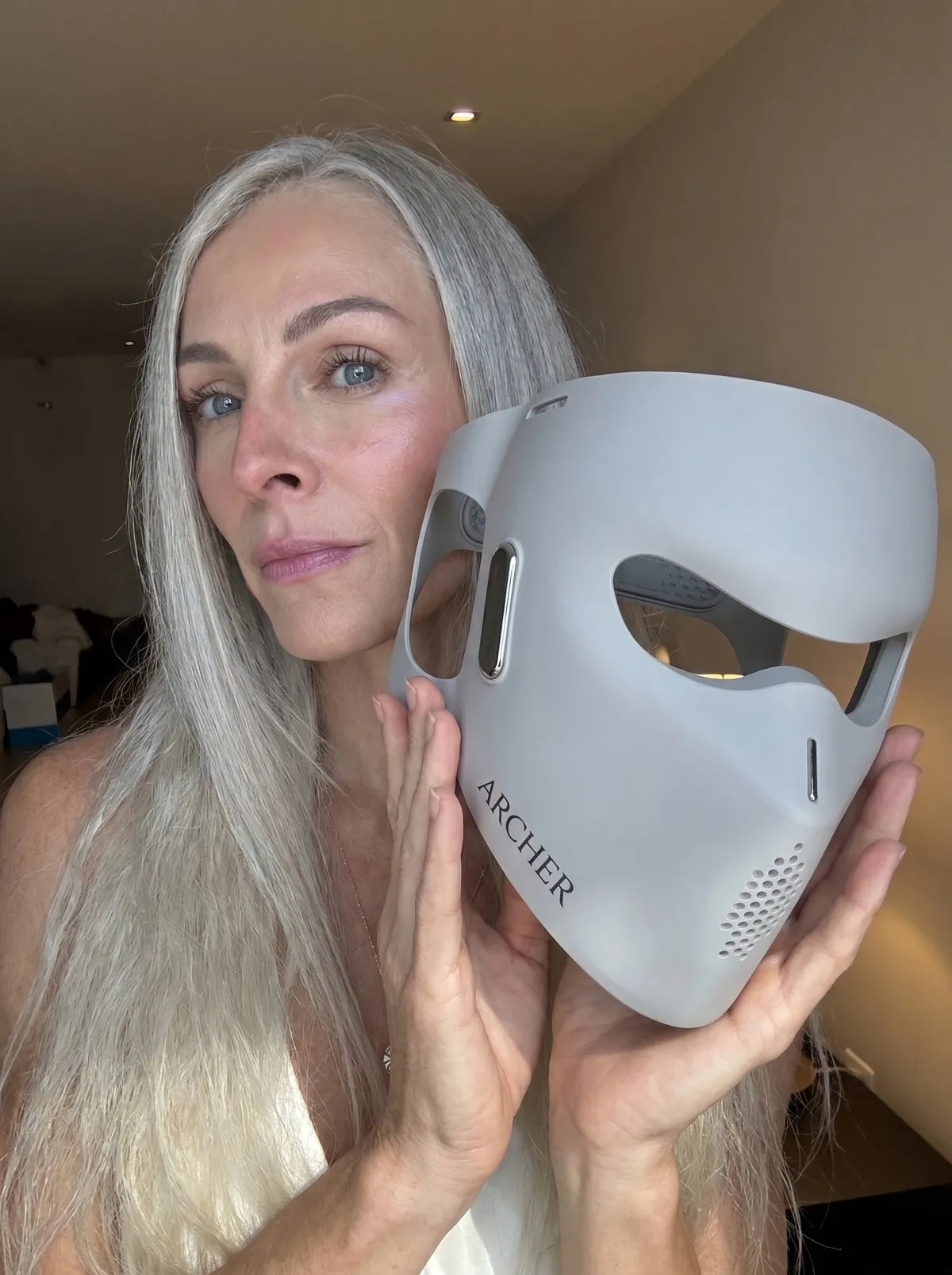 Woman holding the ARCHER LED face mask beside her face in soft natural light.